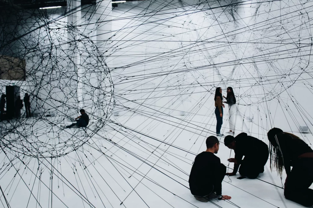 Wide view of Tomás Saraceno's contemporary art installation inside a bright white gallery space at Palais de Tokyo, featuring a vast network of black threads woven into a giant spherical web, with multiple visitors standing and kneeling on the floor to look at the large-scale artwork in Palais de tokyo