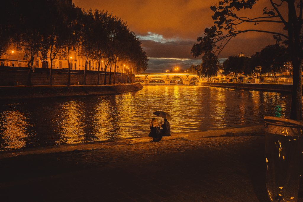 The Seine River by Night in Paris with a couple sitting on the edge by themself holding an umbrella.