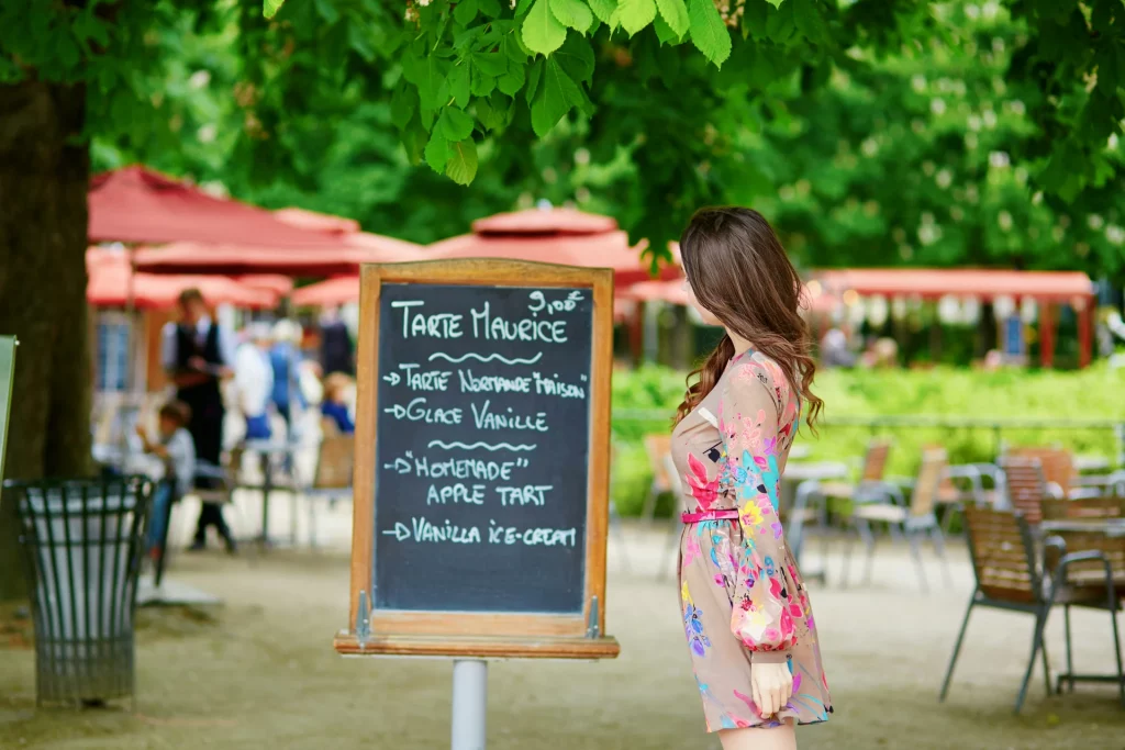 A young Parisian woman reading a handwritten menu at a Parisian restaurant in the Tuileries Garden on a sunny summer day. Learning how to order food in Paris