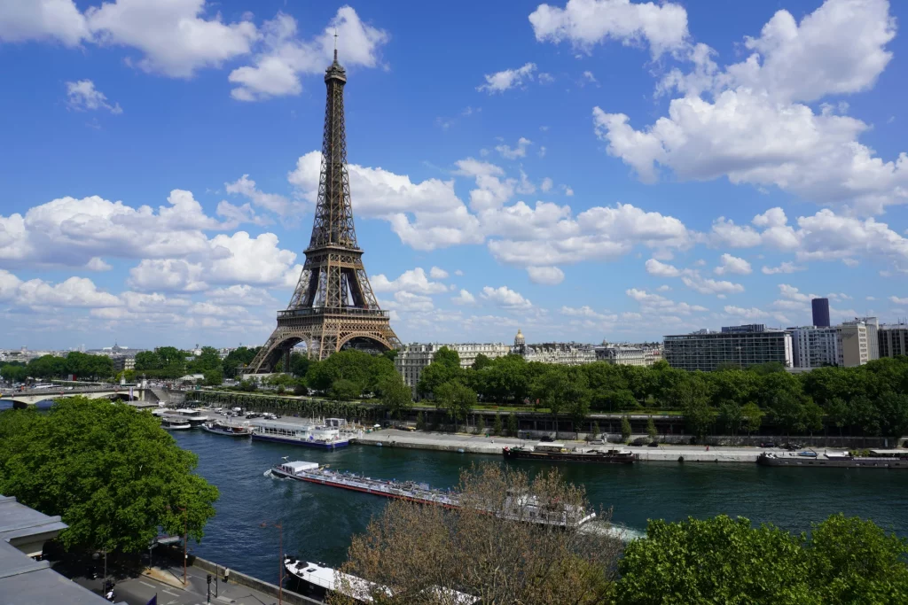 High angle view of the Eiffel Tower rising above green trees under a bright blue sky with white clouds, with a long river boat cruising along the Seine River in the foreground.