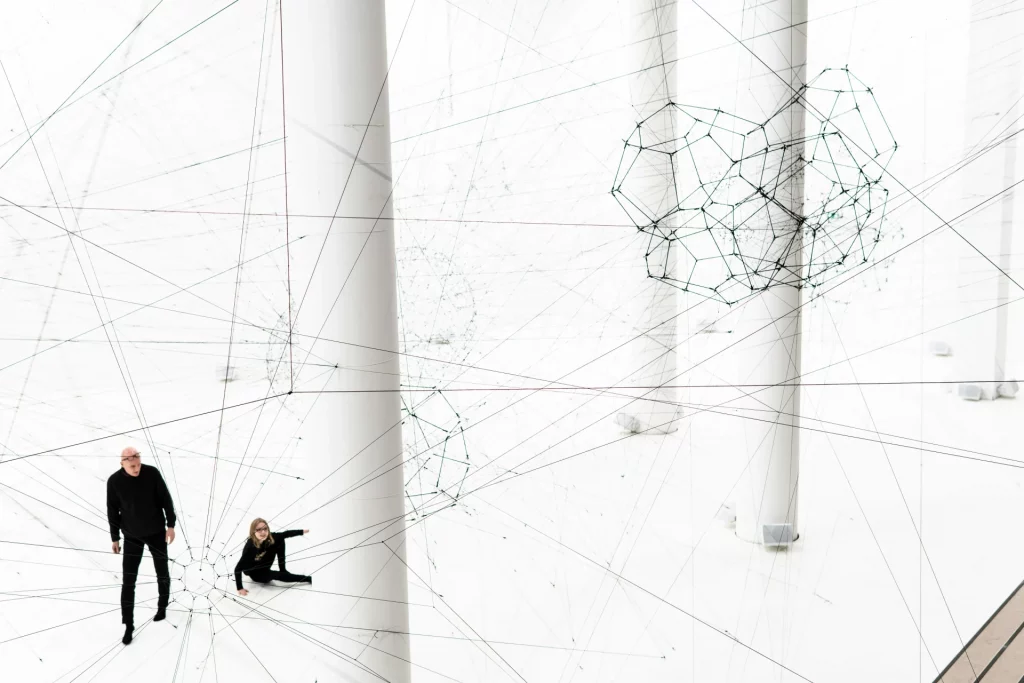 High angle view of Tomás Saraceno's contemporary art installation inside a bright white hall, featuring a massive network of black threads stretched between thick white columns to create giant web structures and floating geometric shapes, with two visitors in black clothing looking at the work on the floor below In Palais de Tokyo Paris
