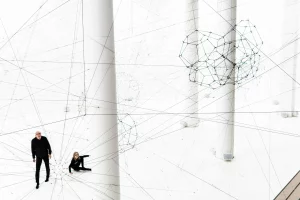 High angle view of Tomás Saraceno's contemporary art installation inside a bright white hall, featuring a massive network of black threads stretched between thick white columns to create giant web structures and floating geometric shapes, with two visitors in black clothing looking at the work on the floor below In Palais de Tokyo Paris