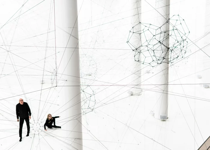 High angle view of Tomás Saraceno's contemporary art installation inside a bright white hall, featuring a massive network of black threads stretched between thick white columns to create giant web structures and floating geometric shapes, with two visitors in black clothing looking at the work on the floor below In Palais de Tokyo Paris
