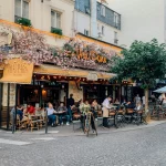 Outdoor view of Le Vrai Paris café in Montmartre, Paris, with customers dining on the terrace beneath pink floral decorations on a warm summer day.
