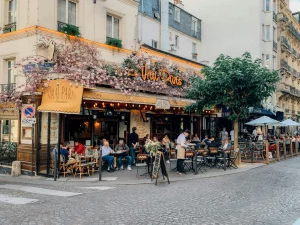 Outdoor view of Le Vrai Paris café in Montmartre, Paris, with customers dining on the terrace beneath pink floral decorations on a warm summer day.
