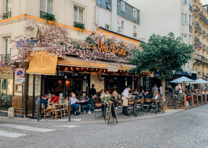 Outdoor view of Le Vrai Paris café in Montmartre, Paris, with customers dining on the terrace beneath pink floral decorations on a warm summer day.