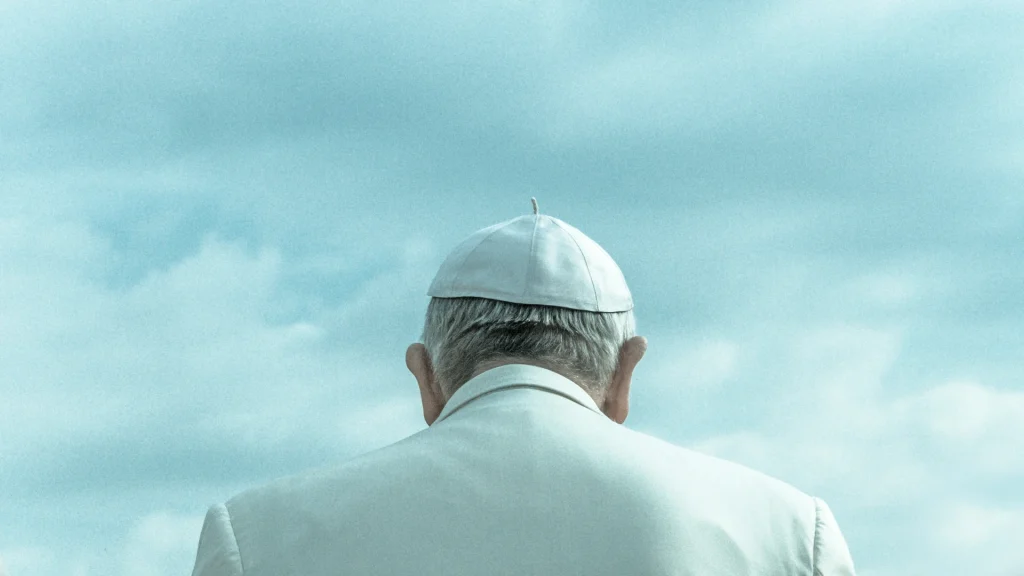 Pope Leo XIV wearing white papal robes and skullcap, viewed from behind against a blue sky