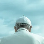 Pope Leo XIV wearing white papal robes and skullcap, viewed from behind against a blue sky
