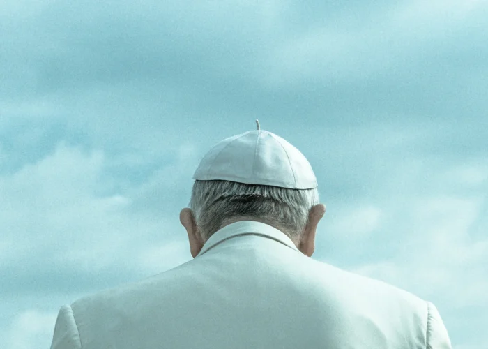 Pope Leo XIV wearing white papal robes and skullcap, viewed from behind against a blue sky