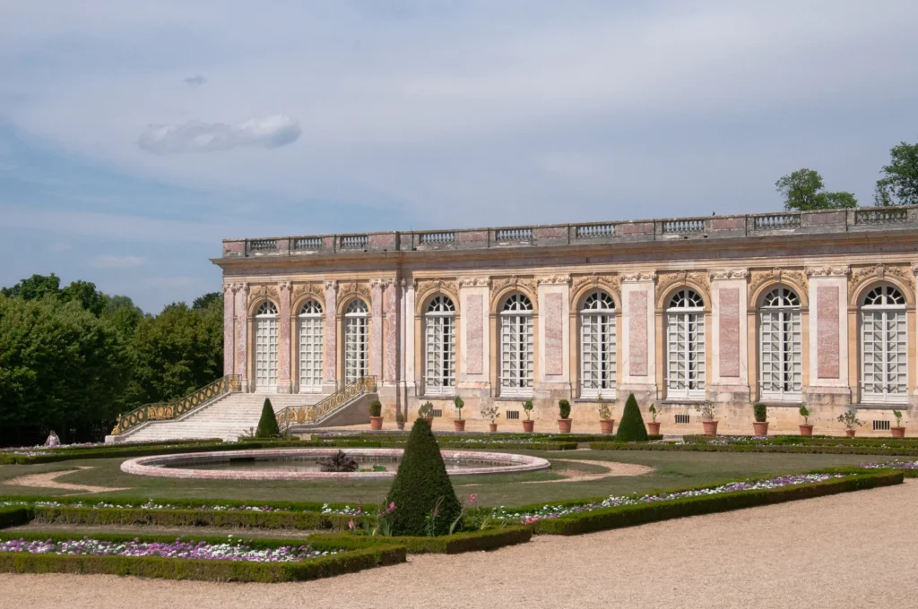 Exterior of The Grand Trianon, overlooking its formal gardens, with ornate pink marble architecture and symmetrical flower beds