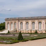 Exterior of The Grand Trianon, overlooking its formal gardens, with ornate pink marble architecture and symmetrical flower beds