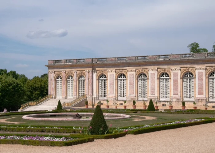 Exterior of The Grand Trianon, overlooking its formal gardens, with ornate pink marble architecture and symmetrical flower beds